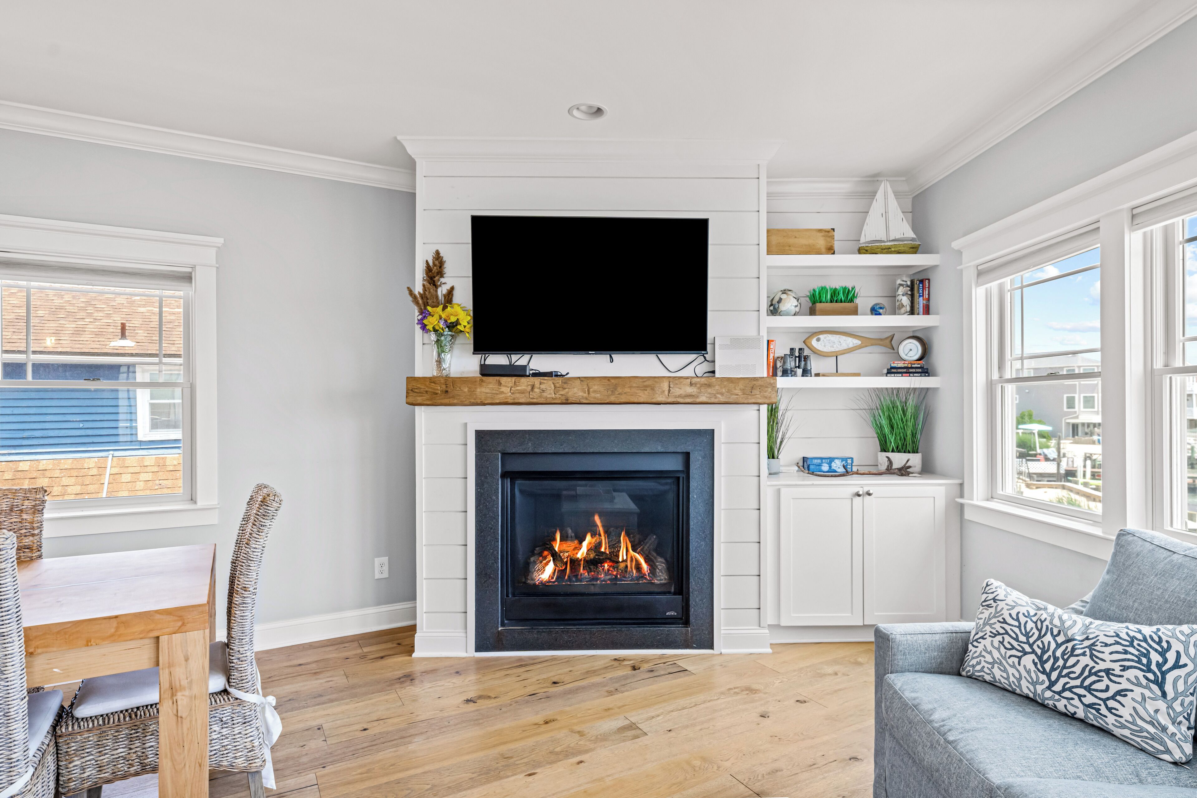 A living room with a fireplace, a flat-screen TV mounted above it, a dining area with wicker chairs, and a view of the outside through large windows.