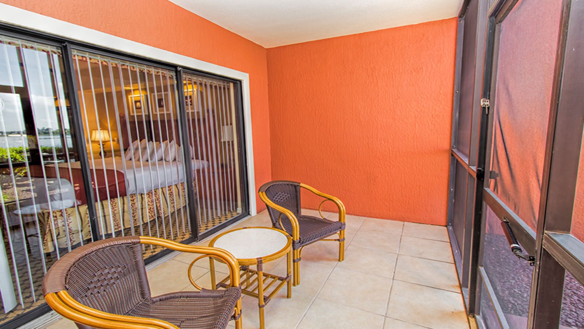 Ein Balkon mit zwei Korbstühlen, einem kleinen Tisch, einer Schiebetür und Blick in einen Raum mit Bett und Vorhängen.