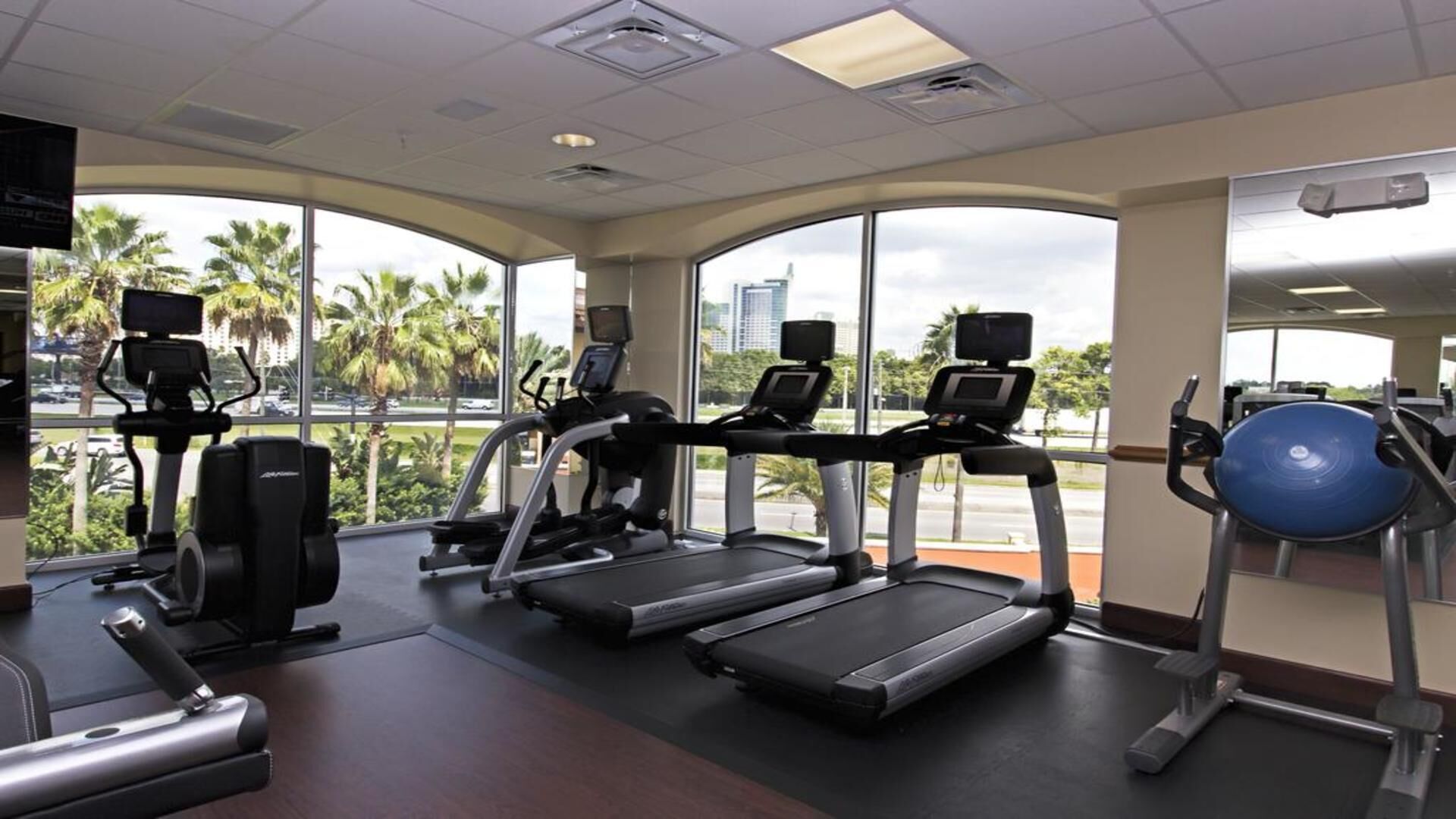 A gym with treadmills, elliptical machines, and a rowing machine, large windows with a city view, and a ceiling with recessed lighting.
