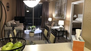Une chambre d’hôtel comprenant un coin repas, deux canapés, un ventilateur de plafond, une grande fenêtre avec des rideaux, un tableau au mur, ainsi qu’une table de chevet avec une lampe et une boîte de mouchoirs.