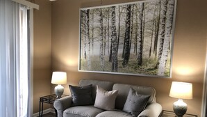 Un salon avec un canapé beige, deux tables d’appoint avec des lampes, une table basse en verre et un grand tableau représentant une forêt, accroché au mur.