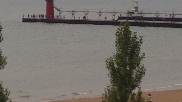 Een vuurtoren op een steiger die uitloopt in kalm water, met een strand en bomen op de voorgrond.