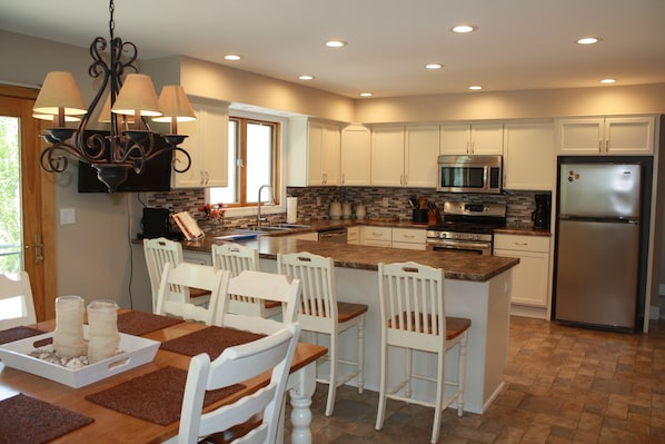 A modern kitchen with white cabinetry, a central island with bar stools, a stainless steel refrigerator, and a chandelier.