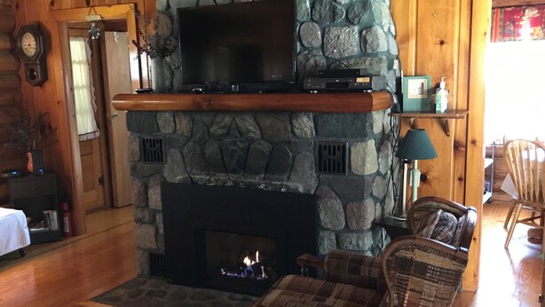 Ein gemütliches Wohnzimmer mit Kamin aus Stein, einem Flachbildfernseher auf einer Holzwand und einem karierten Sofa, sowie ein Essbereich im Hintergrund.