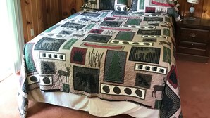 Ein Schlafzimmer mit einer holzverkleideten Wand, einer gesteppten Bettdecke mit Tiermotiven, einem Nachttisch mit Lampe und einem Fenster mit Vorhängen.