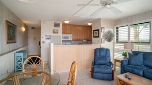 Ein modernes Wohnzimmer mit einem blauen Sofa, einem Holztisch und einem Essbereich mit einem Glastisch und Holzstühlen. Im Hintergrund ist eine Küche mit Holzschränken und einer Mikrowelle erkennbar.