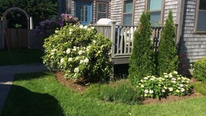 Una casa de dos pisos con una terraza de madera, un pequeño jardín con flores y un césped bien cuidado.