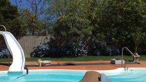 Una piscina con tobogán, una silla de descanso y una valla de madera con flores azules al fondo.
