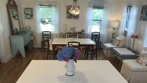 Un comedor con una mesa blanca, sillas y un florero. A la derecha, un banco con cojines y una lámpara.