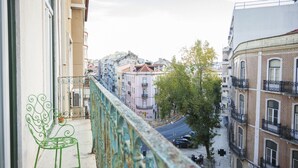 Un balcon avec une chaise en métal vert, donnant sur une rue bordée d’immeubles et de voitures garées.