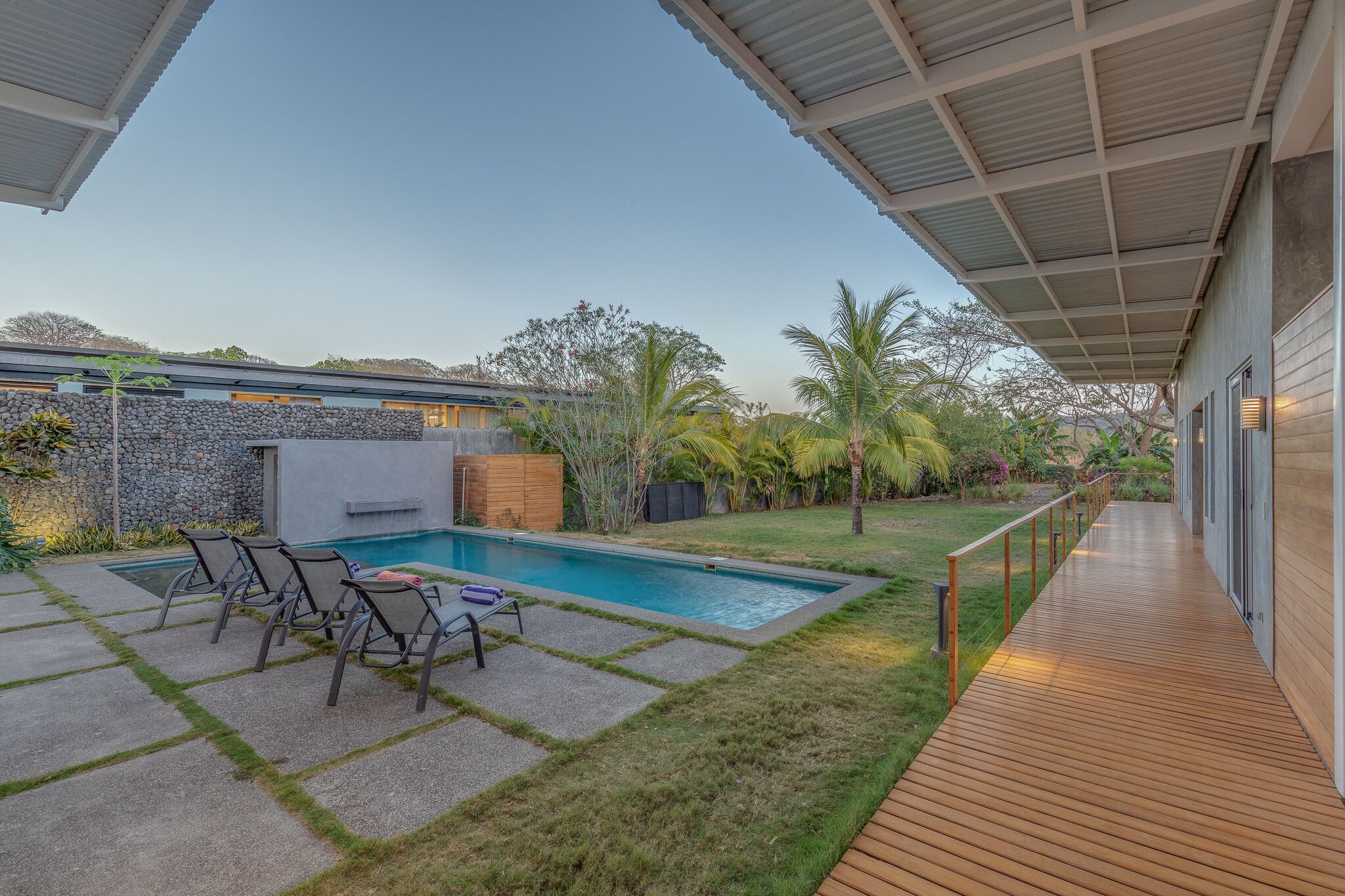 Un área de piscina moderna al aire libre con un deck de madera, sillas de descanso y una pared de piedra.