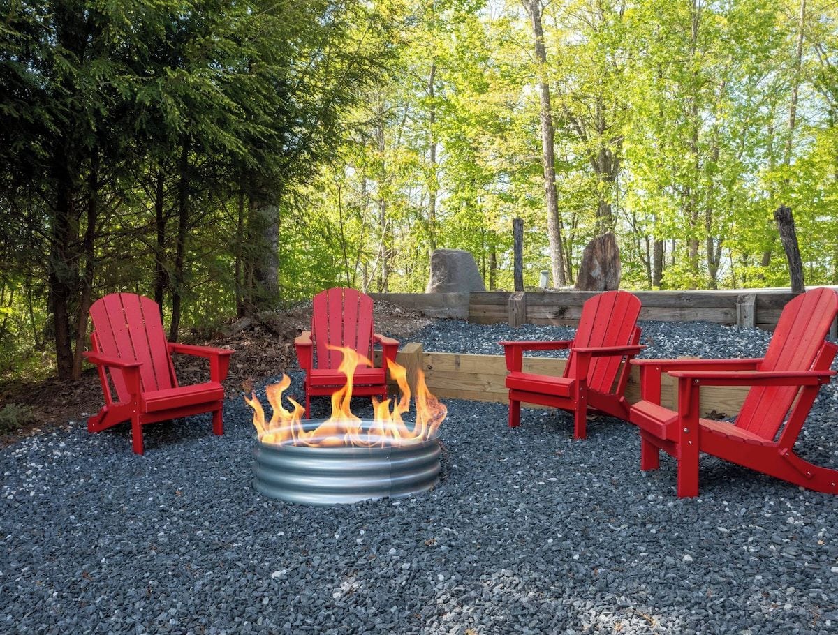A fire pit with four red Adirondack chairs arranged around it on a gravel surface.