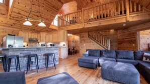 A spacious cabin with a wooden interior, a kitchen area, a seating area with a sofa and ottoman, and a staircase leading to an upper level. - Heavenly Mtn Views, Hot Tub+Fire Pit+Grill, Fireplace, WiFi, Hike, Close to BRP! (Spruce Pine)