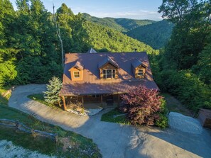 A two-story wooden cabin with a covered porch, surrounded by dense forest and hills. - Heavenly Views~Hot Tub+Fire Pit+Fireplace & WiFi! (Spruce Pine)