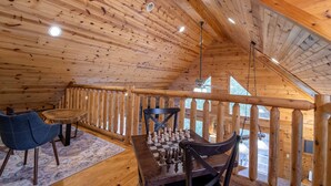 A wooden interior with a chessboard on a table, a blue chair, and a wooden railing. - Heavenly Mtn Views, Hot Tub+Fire Pit+Grill, Fireplace, WiFi, Hike, Close to BRP! (Spruce Pine)