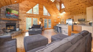 A spacious living area with a fireplace, large windows, and a kitchen area. - Heavenly Mtn Views, Hot Tub+Fire Pit+Grill, Fireplace, WiFi, Hike, Close to BRP! (Spruce Pine)