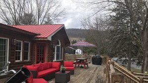 Eine Holzterrasse mit einer blockhafenähnlichen Hütte mit rotem Dach, Sitzgelegenheiten im Freien und Blick auf einen See.