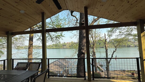 A covered deck with a table and chairs, overlooking a lake with trees and buildings in the background.