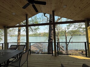 Una terraza techada con una mesa y sillas, con vista a un lago rodeado de árboles y edificios al fondo.