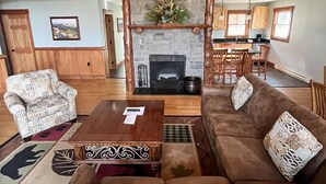 A cozy living room with a stone fireplace, wooden ceiling, and a chandelier. There is a sofa set and a coffee table with a remote control on it.