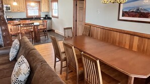 A spacious living area with a brown leather sofa, wooden dining table, and chairs. A chandelier hangs above the table.