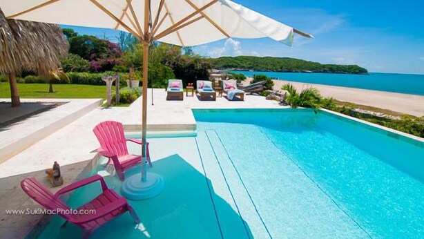 Ein Schwimmbecken mit Liegestühlen und Sonnenschirmen, Blick auf einen Strand und eine tropische Gartenanlage.