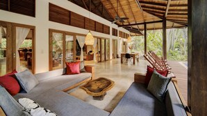 A spacious living area with a high ceiling, wooden beams, and large windows. There are multiple seating areas with sofas and a coffee table.