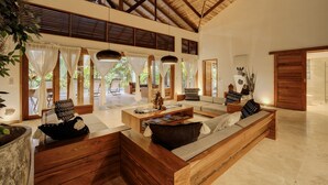 A spacious living area with a high ceiling, wooden furniture, and large windows allowing natural light.