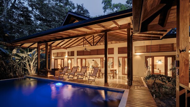 A modern outdoor pool area with wooden deck and lounge chairs, surrounded by a covered patio with large windows.