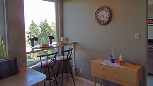Een keuken met een eethoek, een houten tafel, twee barkrukken, een klok en een raam met uitzicht op bomen.