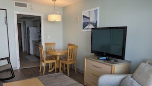 En stue med fjernsyn, spisebord med stole, sofa og et køkken i baggrunden.