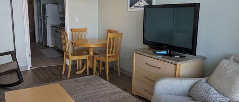 A living room with a television, a dining table with chairs, a sofa, and a kitchen in the background.