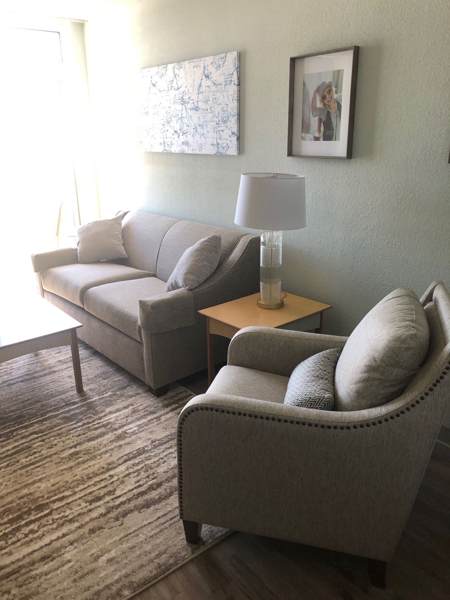 A living room with a sofa, armchair, and a small table with a lamp. There is a framed picture on the wall and a rug on the floor.