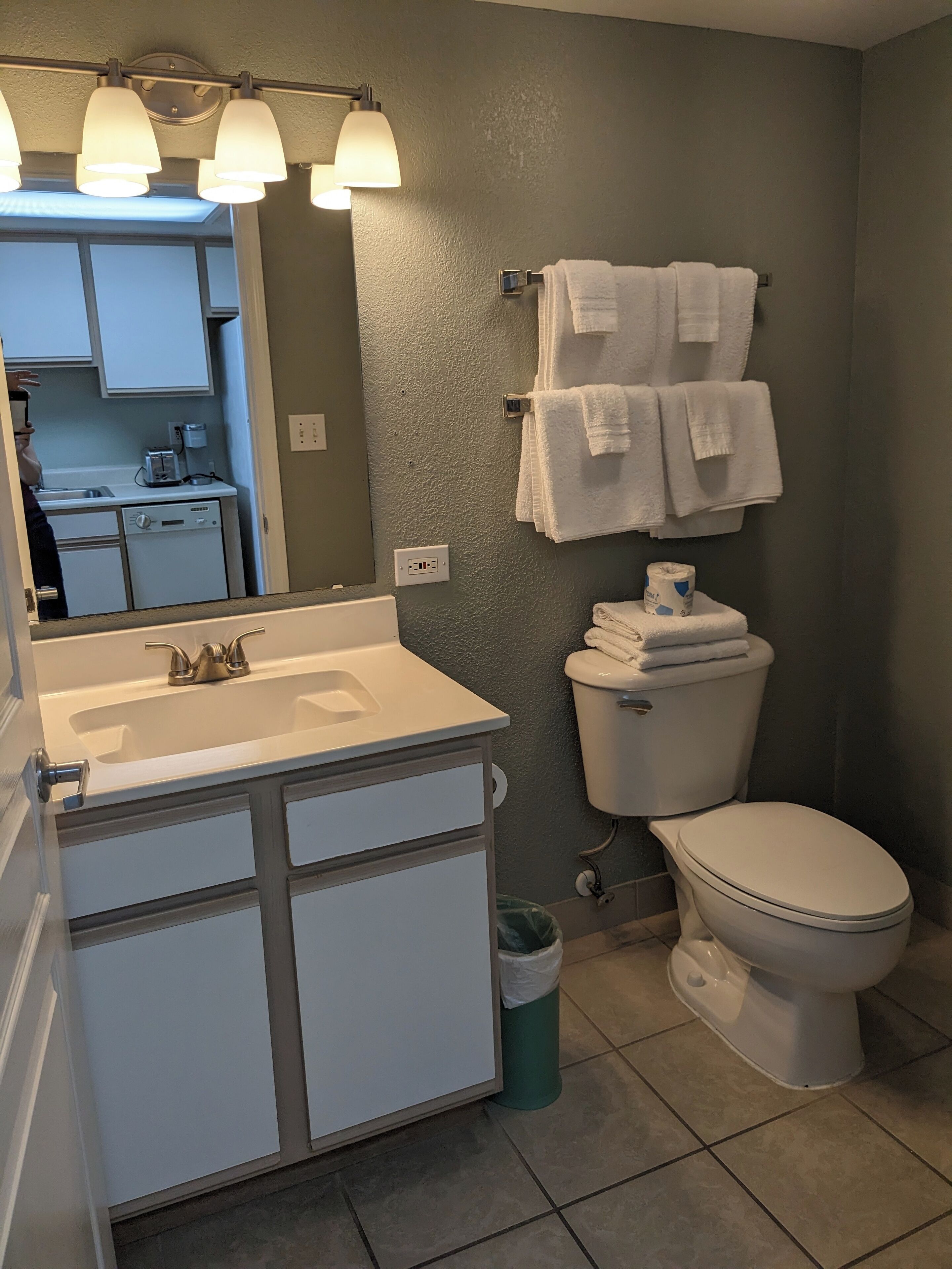 Une salle de bain avec un lavabo blanc, des toilettes et un miroir avec éclairage. Des serviettes sont accrochées au mur, et il y a une poubelle.