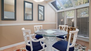 Un comedor con una mesa de vidrio rodeada de sillas blancas con cojines azules, paredes color beige y un tragaluz.