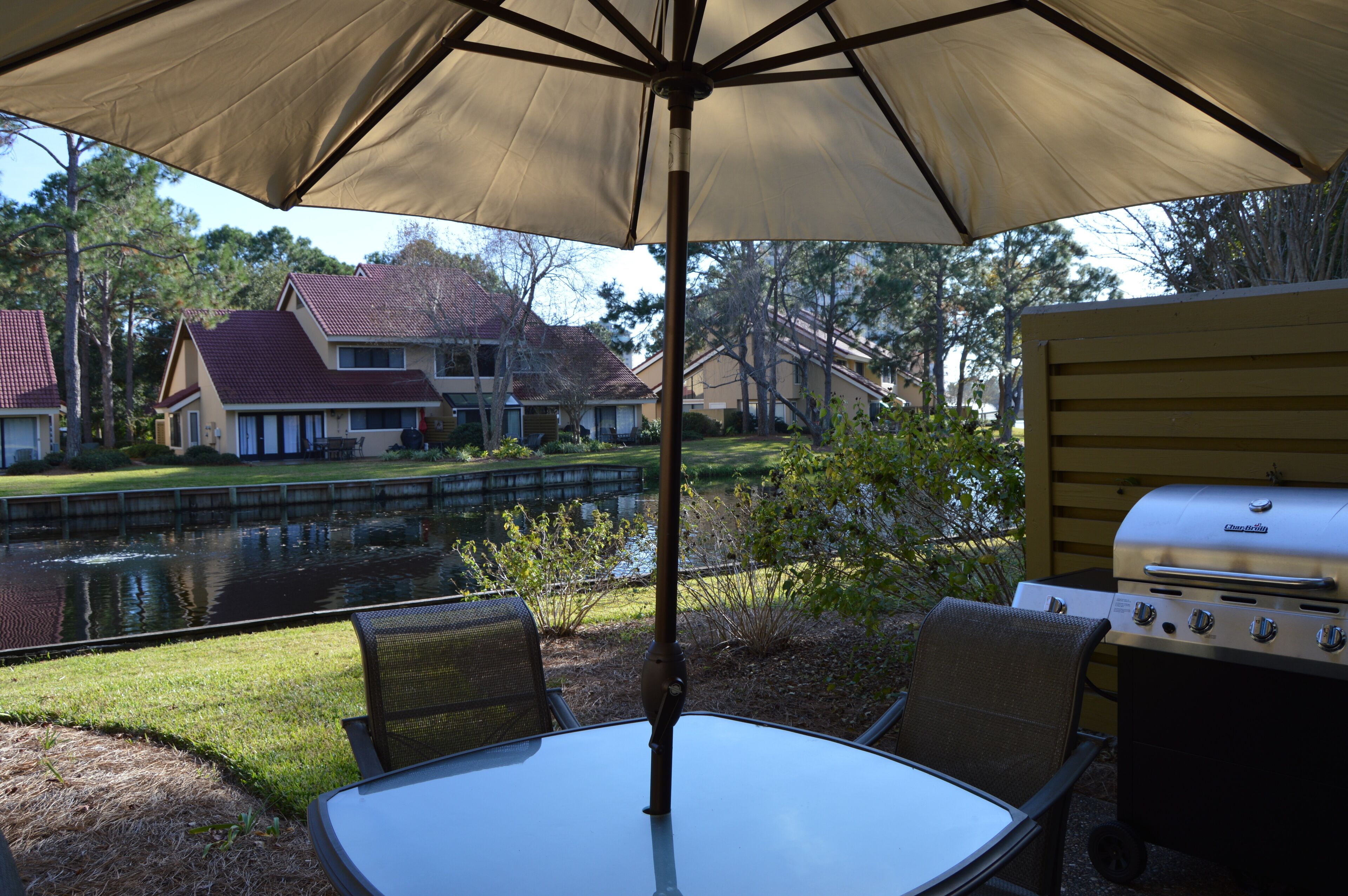 An outdoor patio with a table and chairs under a large umbrella, a grill, and a view of houses and a pond.