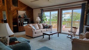 A spacious living room with a beige sofa, a blue sofa, and a wooden rocking chair. A coffee table is in the center, and there is a large window with a view of a balcony and houses. - "He Crab She Crab"  Waterfront Home w/Beach, Bay&Backyard Fun. Perfect for Kids! (Virginia Beach)