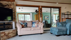 A living room with a beige sofa, a blue armchair, a wooden coffee table, and a TV on a brick wall. - "He Crab She Crab"  Waterfront Home w/Beach, Bay&Backyard Fun. Perfect for Kids! (Virginia Beach)