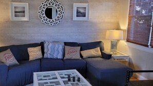 Una sala de estar con un sofá azul oscuro, tres almohadas estampadas, una mesa de centro con una revista, dos mesas auxiliares con lámparas y un espejo decorativo en la pared.