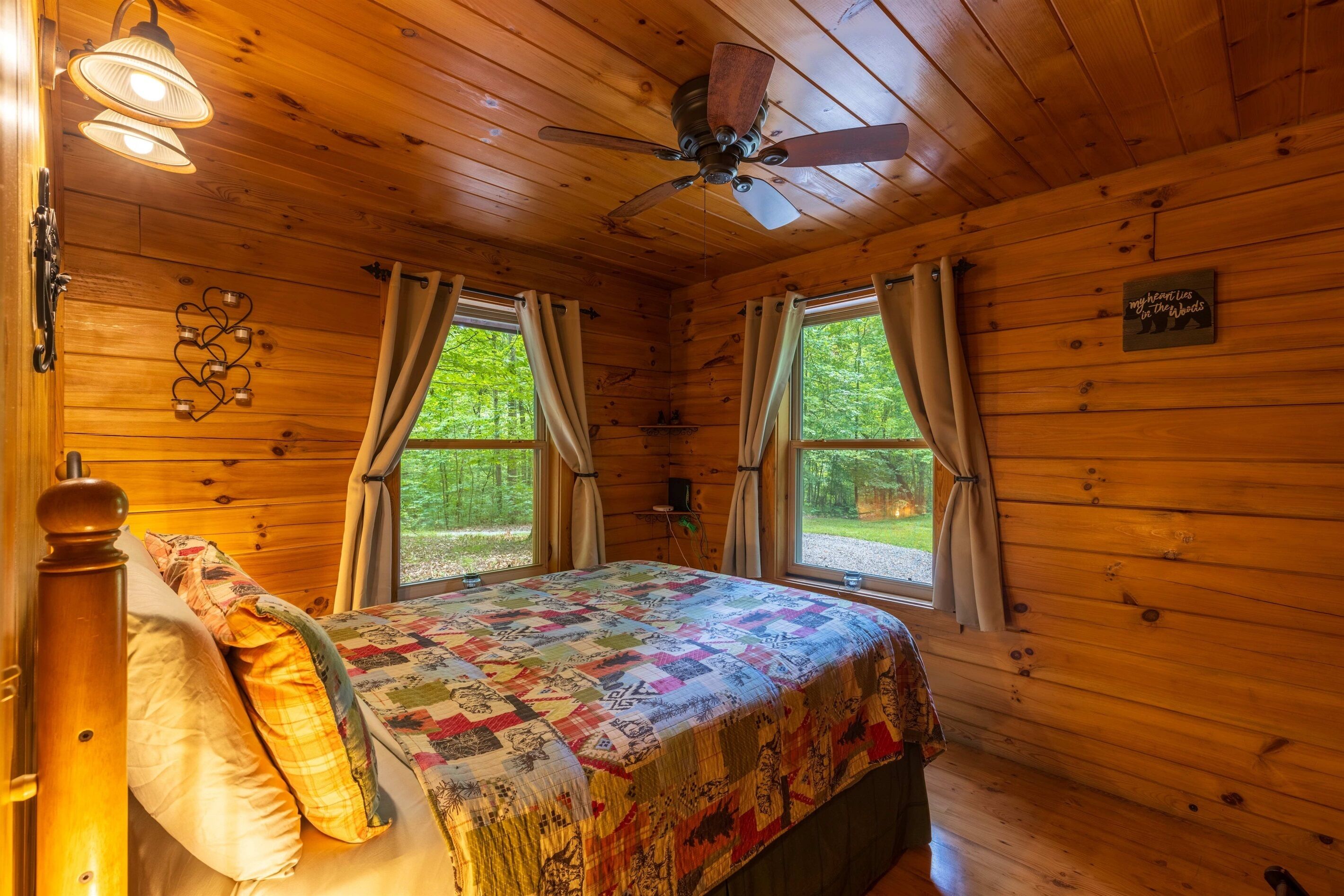 Un dormitorio de cabaña acogedor con techo de madera, cama con colcha colorida, dos ventanas con cortinas y un ventilador de techo.