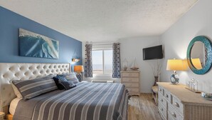 A bedroom with a tufted headboard, a large bed with striped bedding, a television, a mirror, and a dresser with drawers. - Oceanfront Condo in High Rise in the Heart of the Resort Area (Virginia Beach)