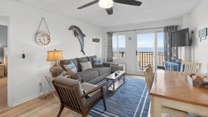 A living room with a ceiling fan, a sofa, a coffee table, and a dining table. - Oceanfront Condo in High Rise in the Heart of the Resort Area (Virginia Beach)