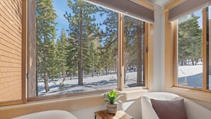 Een kamer met een groot raam dat uitzicht geeft op een besneeuwd landschap en hoge bomen. Er staat een klein houten tafeltje met een potplant en een kussen erop.