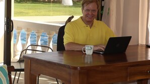 Ein Mann sitzt an einem Holztisch mit einem Laptop und einer Tasse, in einem Zimmer mit Blick auf einen Garten und ein Schwimmbad.