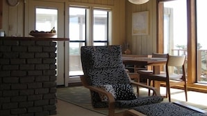 A cozy room with a wooden ceiling, a brick fireplace, a patterned armchair, and a dining area with wooden furniture. - Seaside Modern - Easy 3 min Walk to Beach! (Dillon Beach)