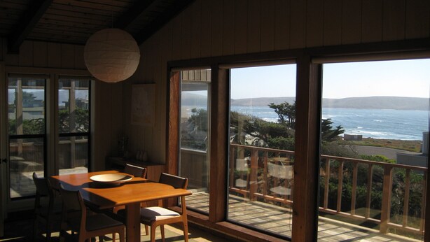 A room with a wooden table and chairs, large windows with a view of the ocean, and a hanging light fixture. - Seaside Modern - Easy 3 min Walk to Beach! (Dillon Beach)