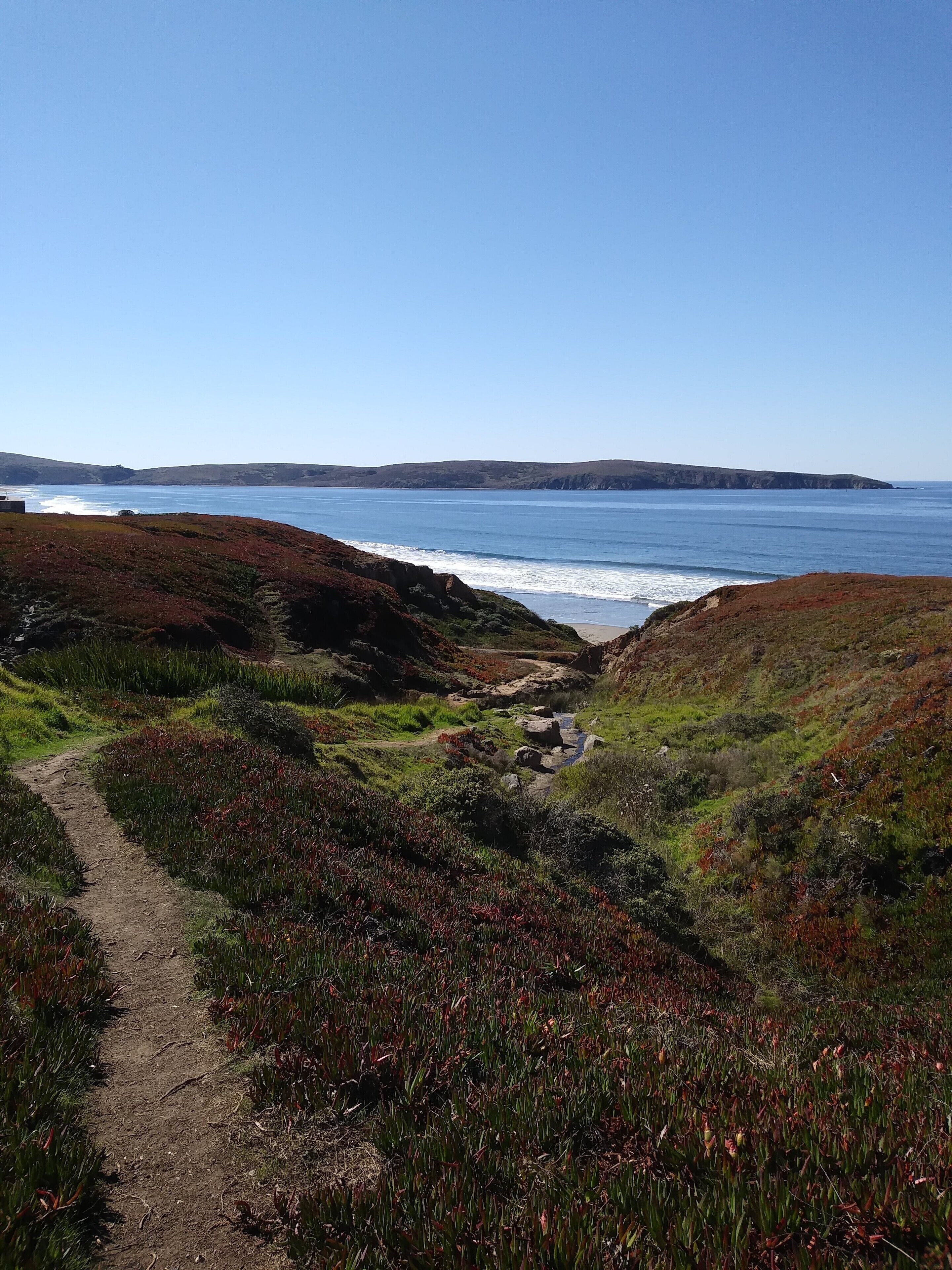 A coastal path leading to a beach with a clear view of the ocean and distant hills.
