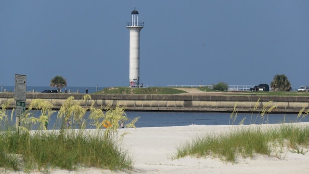 Un phare au bord de l’eau, avec une plage de sable et des dunes herbeuses au premier plan.