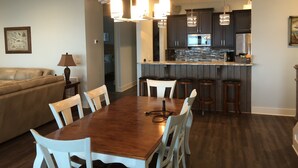 Un comedor con mesa de madera y sillas blancas, una cocina con armarios oscuros y electrodomésticos de acero inoxidable, y una sala de estar con sofá beige y una lámpara.