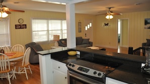 A modern kitchen with a black countertop, white cabinetry, and a built-in oven. There is a dining area with a wooden table and chairs, and a living room with a gray couch and a coffee table. - Steps to the beach.  Pet friendly Beach House w/pool!  4 Bedrooms sleeps 12! (North Myrtle Beach)